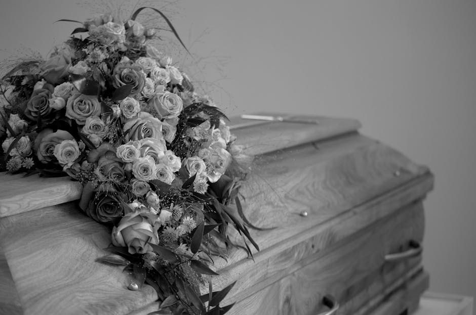 A detailed black-and-white photograph of a large floral arrangement placed atop a closed wooden casket. The bouquet consists of numerous roses in various shades, arranged with lush greenery and delica