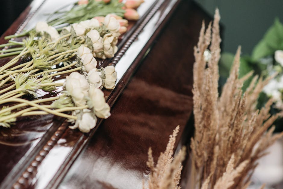 A close-up of a floral arrangement on a polished wooden surface, featuring a bouquet of white and pale pink roses with green stems and leaves, wrapped in delicate tissue paper. To the right, tall beig