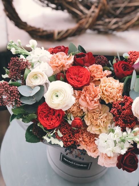 A round floral gift arrangement featuring a variety of fresh flowers including deep red roses, soft peach carnations, and white ranunculi, complemented by sprigs of green eucalyptus and dark red berri
