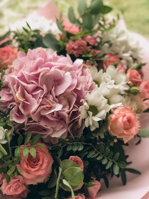 A close-up image of a floral arrangement featuring large, blush pink hydrangea blooms with ruffled petals, complemented by smaller peach roses and white daisy-like flowers with yellow centers. The bou