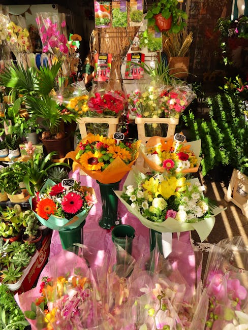 A vibrant display of fresh floral bouquets arranged on a pink cloth-covered table inside a flower shop. The bouquets feature bright, colorful flowers such as yellow sunflowers, pink gerbera daisies, a