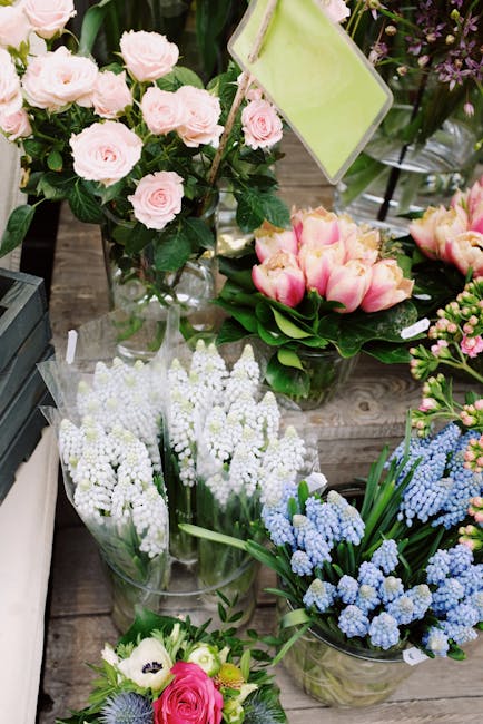 A selection of freshly cut flowers arranged in clear glass vases and bouquets, displayed on a rustic wooden surface. The arrangement includes pale pink roses with lush green leaves, delicate white lil